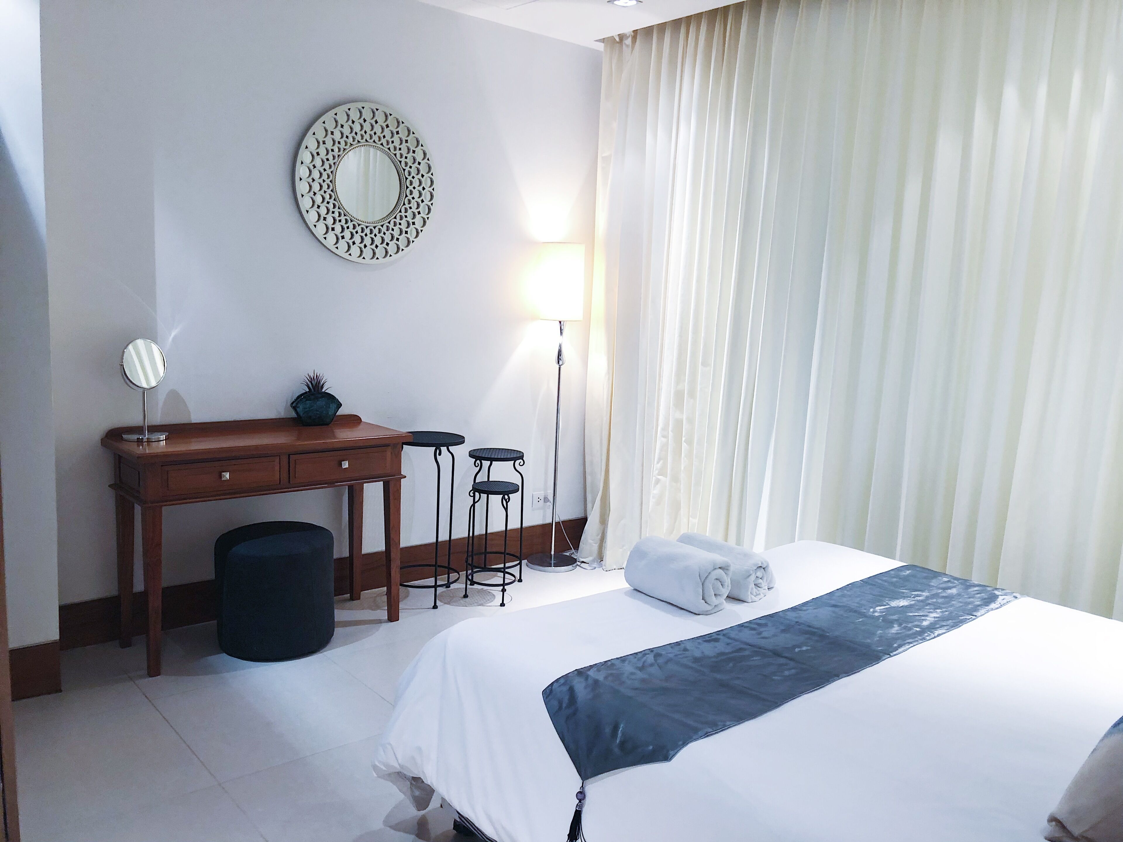 A hotel room with a bed, a desk, a chair, a small table, a trash bin, a lamp, and a window with curtains.