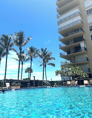 A swimming pool with clear blue water, surrounded by palm trees and a multi-story building with balconies. - Oceanfront 2 bedroom Condo on Hutchinson Island (Fort Pierce)