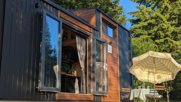 Una cabaña moderna con fachada de madera, puerta corrediza de vidrio y un pequeño espacio exterior con sombrilla.