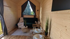 Interior acogedor de una cabaña con una mesa de comedor para dos, un sofá verde y una ventana con cortinas.