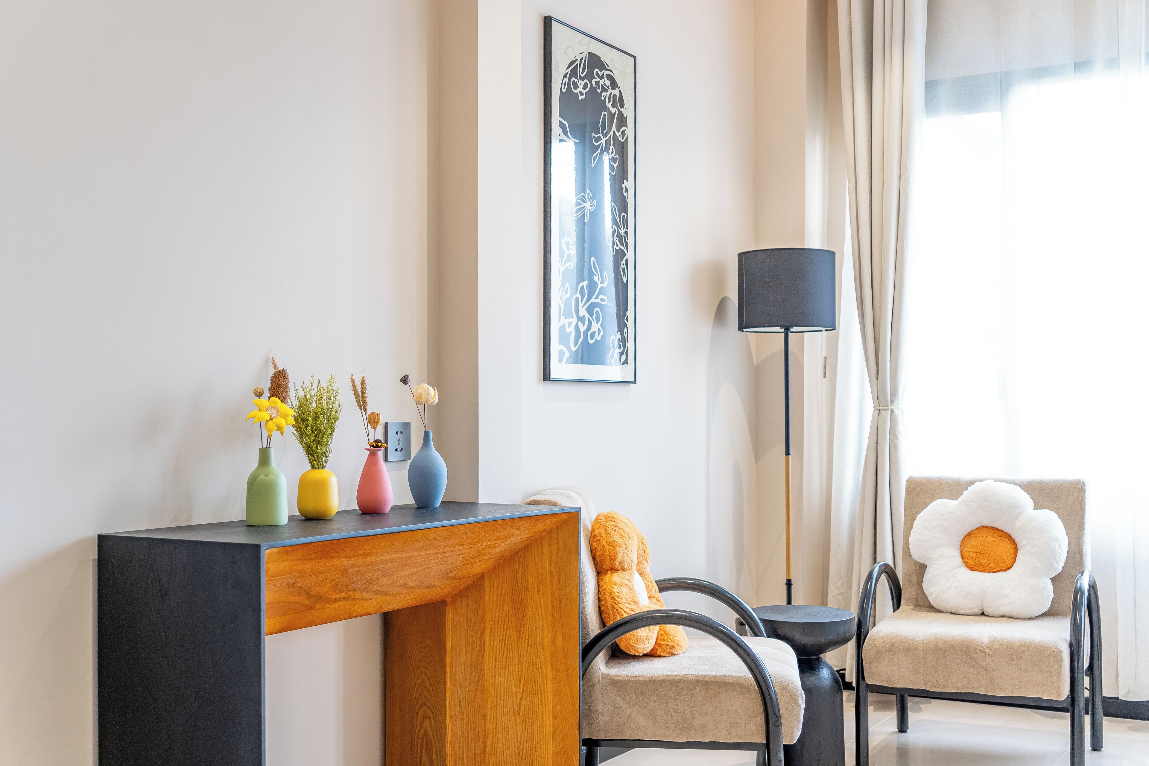 Une chambre d’hôtel moderne avec un bureau en bois, un canapé agrémenté d’un coussin en forme de fleur et une chaise avec un coussin similaire. On trouve trois vases avec des fleurs sur le bureau, ainsi qu’une œuvre d’art encadrée au mur.