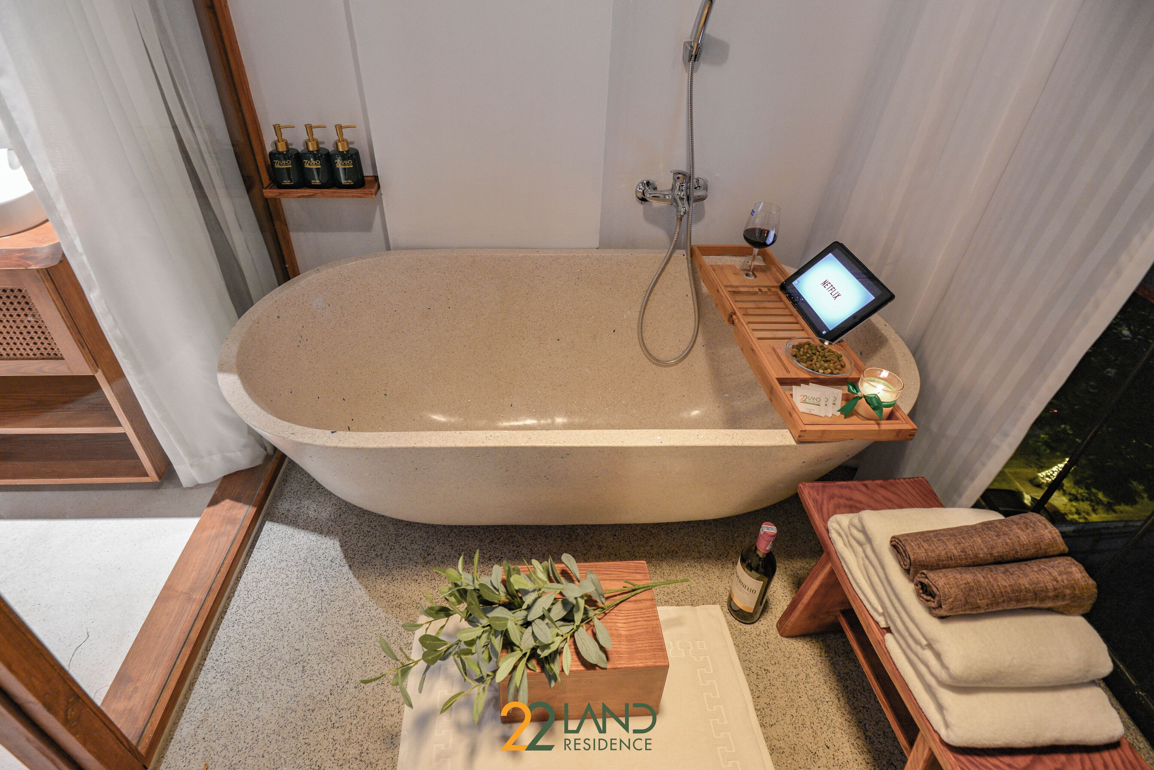 Un baño moderno con una bañera independiente, un estante de madera con artículos de aseo, una mesita con una copa de vino y una tableta, y un banco de madera con toallas.