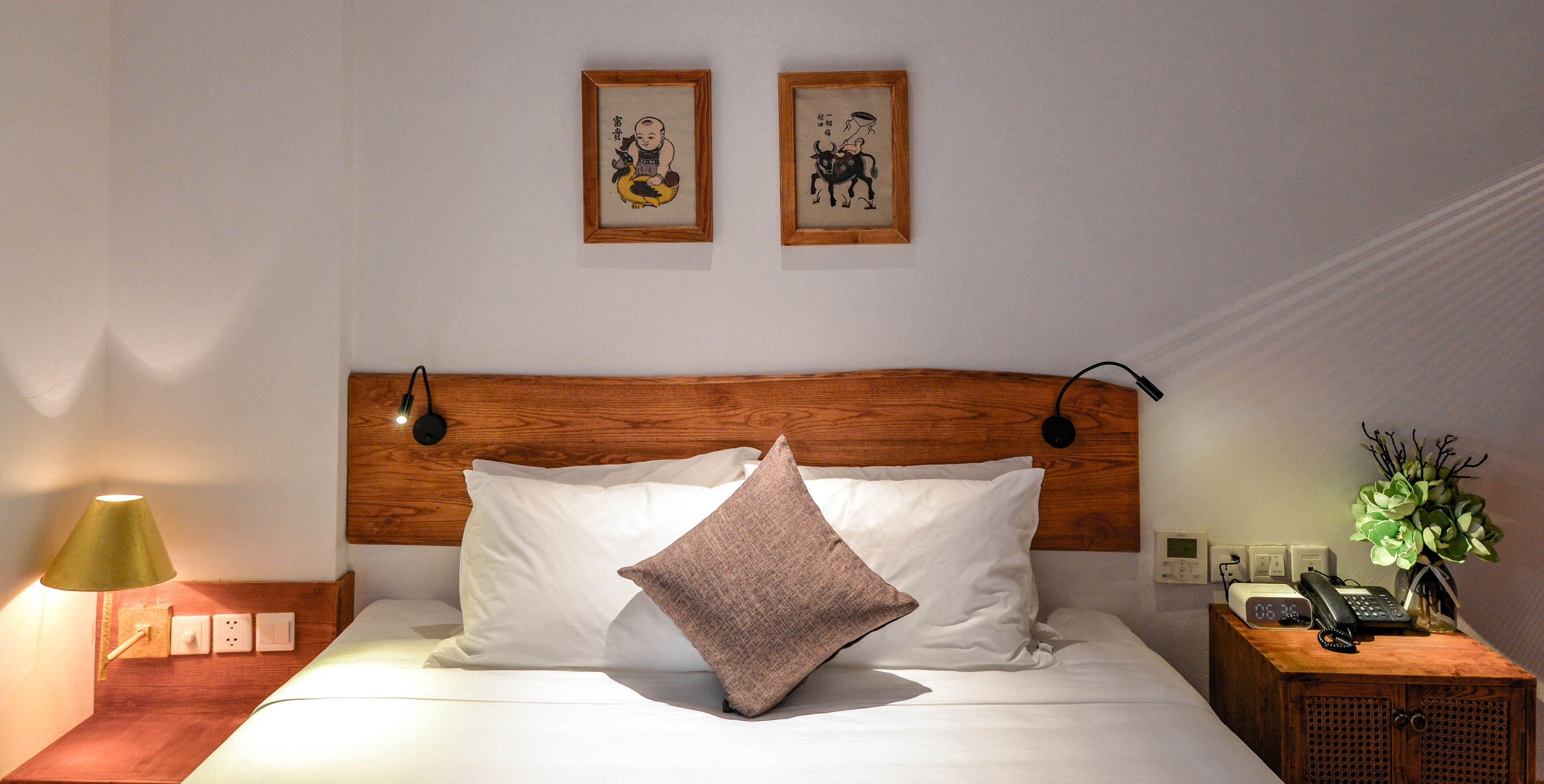 Una cama bien hecha con almohadas blancas y marrones, un cabecero de madera, una lámpara de noche y una mesita de noche con un teléfono y una planta.