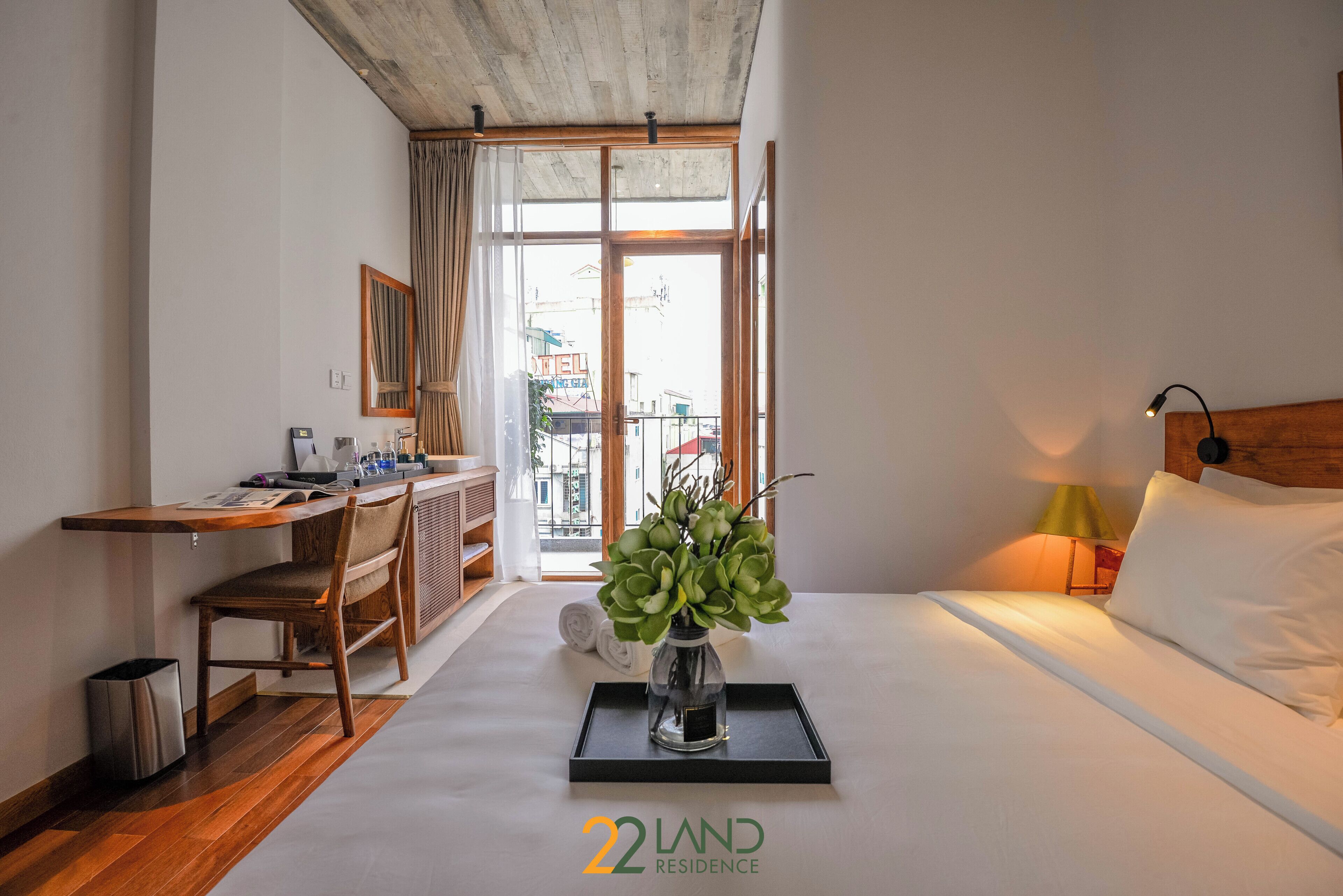 Une chambre d’hôtel avec un lit, un bureau, une chaise, une petite table ornée d’un vase de fleurs et une vue sur la ville par la fenêtre.