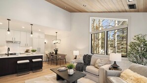 A modern kitchen with white cabinets, a dining area, and a large window with a view of trees.