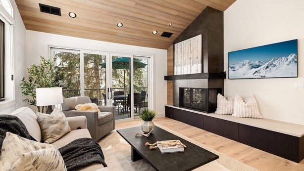 A modern living room with a fireplace, a sofa, a coffee table, and a large window overlooking a deck.