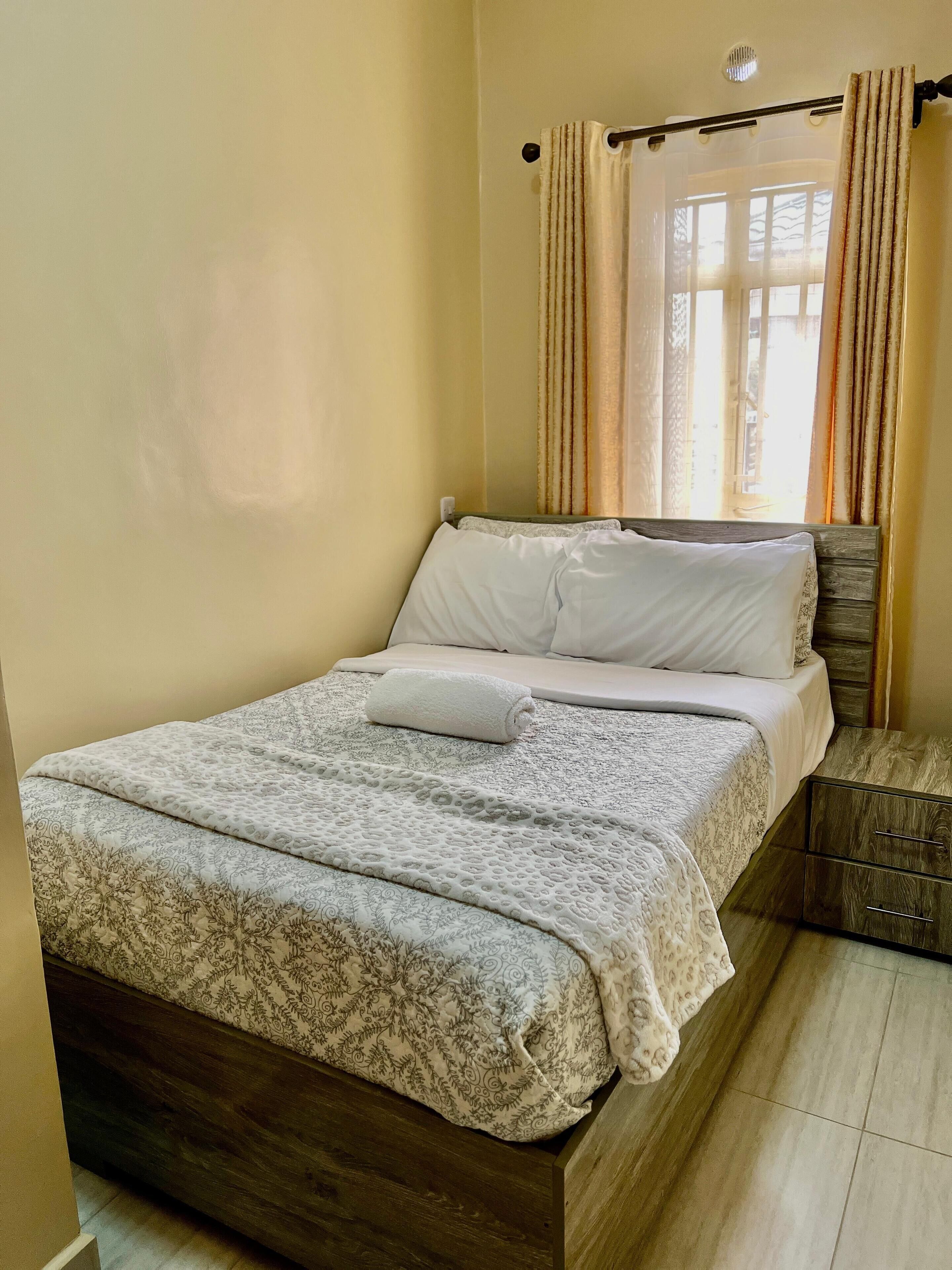 A neatly made bed with a patterned bedspread and pillows, a wooden nightstand with a drawer, and a window with curtains.