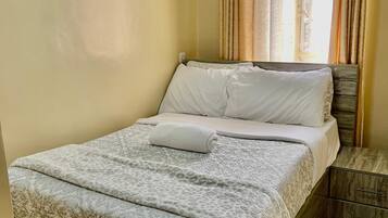 A neatly made bed with a patterned bedspread and pillows, a wooden nightstand with a drawer, and a window with curtains.