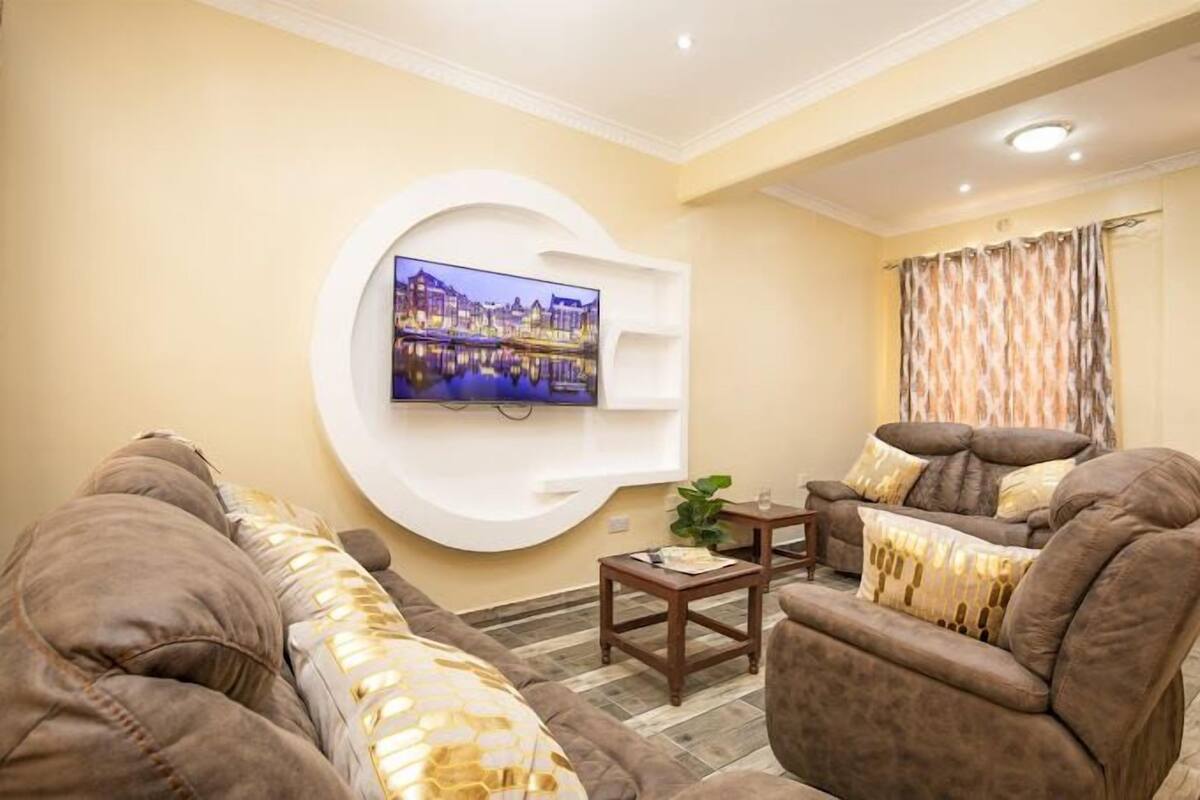A living room with a flat-screen TV mounted on a white wall, a brown sofa, a wooden coffee table, and a patterned curtain.