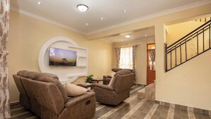 A living room with a flat-screen TV mounted on the wall, two reclining sofas, a small coffee table, and a staircase leading to another level. - Lux Suites Sunset Delight Holiday Homes (Naivasha)
