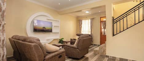 A living room with a flat-screen TV mounted on the wall, two reclining sofas, a small coffee table, and a staircase leading to another level.
