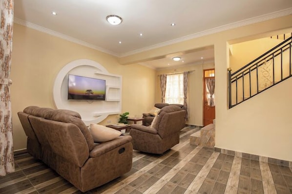 Una sala de estar con un televisor de pantalla plana montado en la pared, dos sillones reclinables, una pequeña mesa de centro y una escalera que conduce a otro nivel.