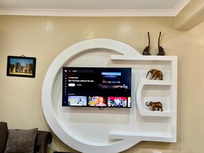 Un salon avec un grand téléviseur à écran plat fixé à un mur blanc, un tableau encadré représentant des éléphants et une étagère blanche sur laquelle sont posées deux statuettes d’éléphants.