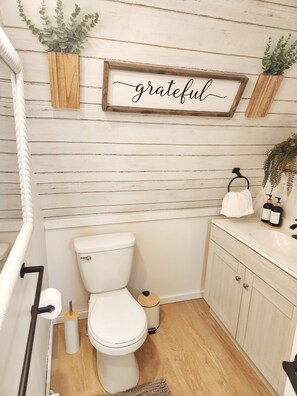 A bathroom with a white toilet, wooden floor, and a 'grateful' sign on the wall. - Magic Mushroom A-Frame Cabin with wood-burning hottub (Sunshine Valley)