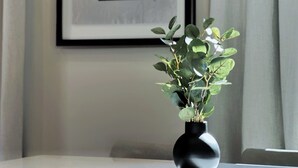 Une œuvre d’art abstraite encadrée accrochée au mur, un vase noir orné de feuillage vert posé sur une table, et un rideau partiellement tiré sur le côté.