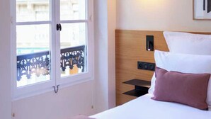 A hotel room with a bed, a nightstand, a wall-mounted light, and a window with a view of buildings. - Hotel Tinah: Elegance and Comfort Near the Champs-elysees (Paris)