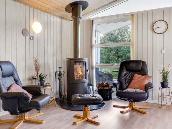 Een gezellige woonkamer met een houtkachel, twee leren fauteuils, een rond hockertje en een groot raam met uitzicht op groen.