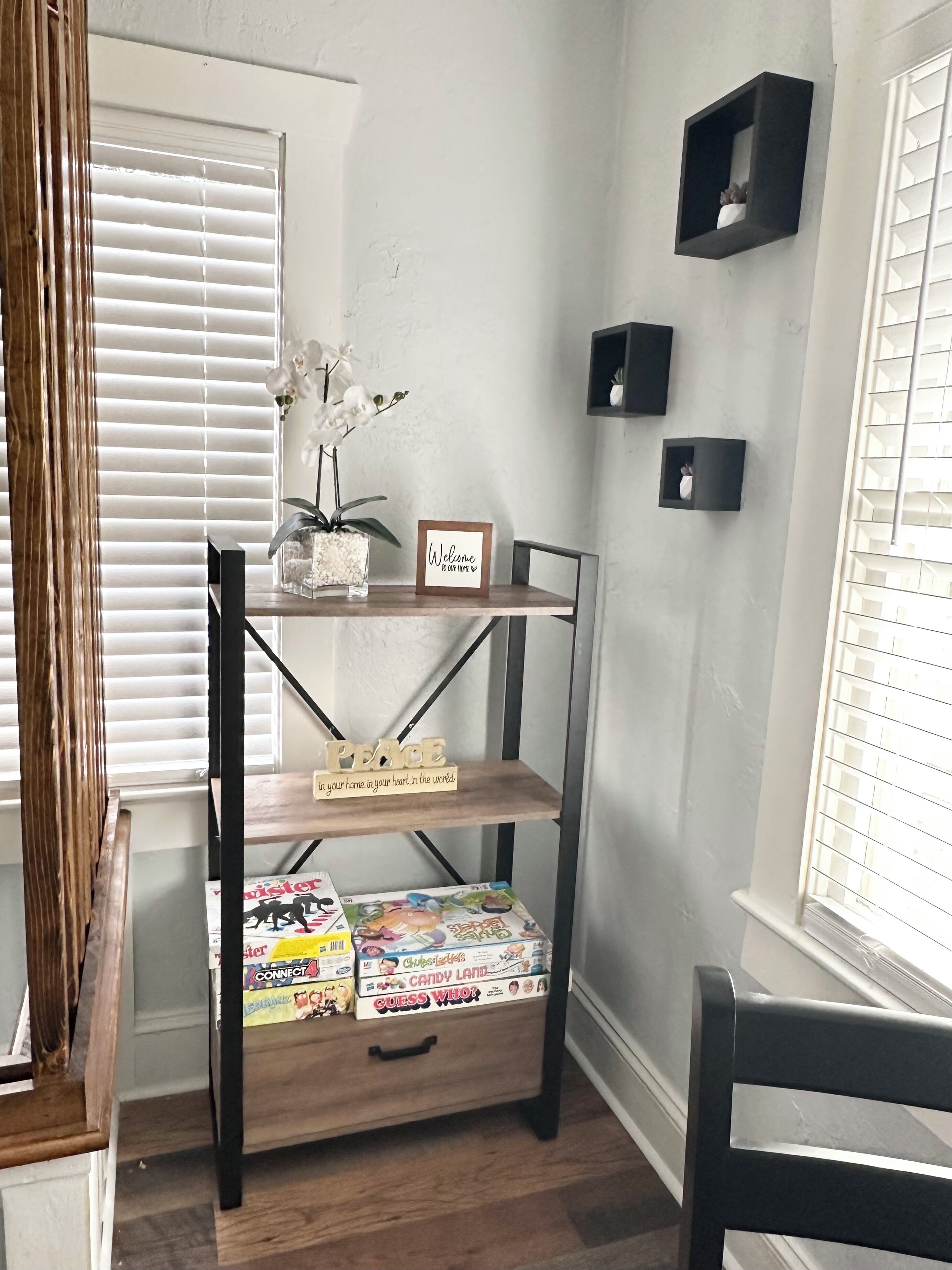 A room with a wooden shelf unit displaying books and a small potted orchid. There are two black wall-mounted shelves, one with a plant and the other empty. A window with blinds is visible, and a chair is positioned near the window.