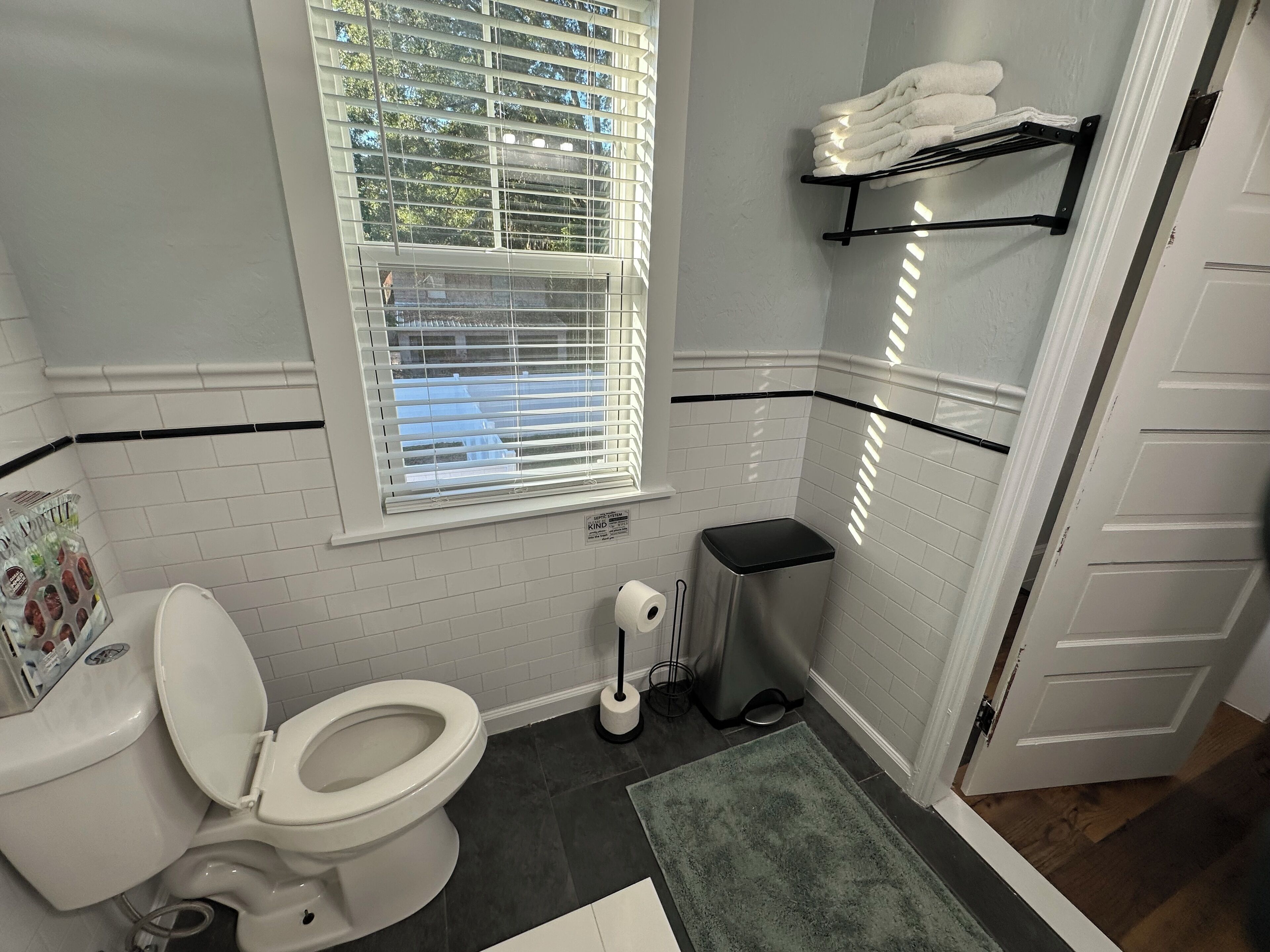 Un baño con inodoro, un cubo de basura, una ventana con persianas y un toallero.