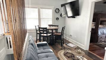 A modern living room with a grey sectional sofa, wooden floor, ceiling fan, dining table with chairs, and a TV mounted on the wall.