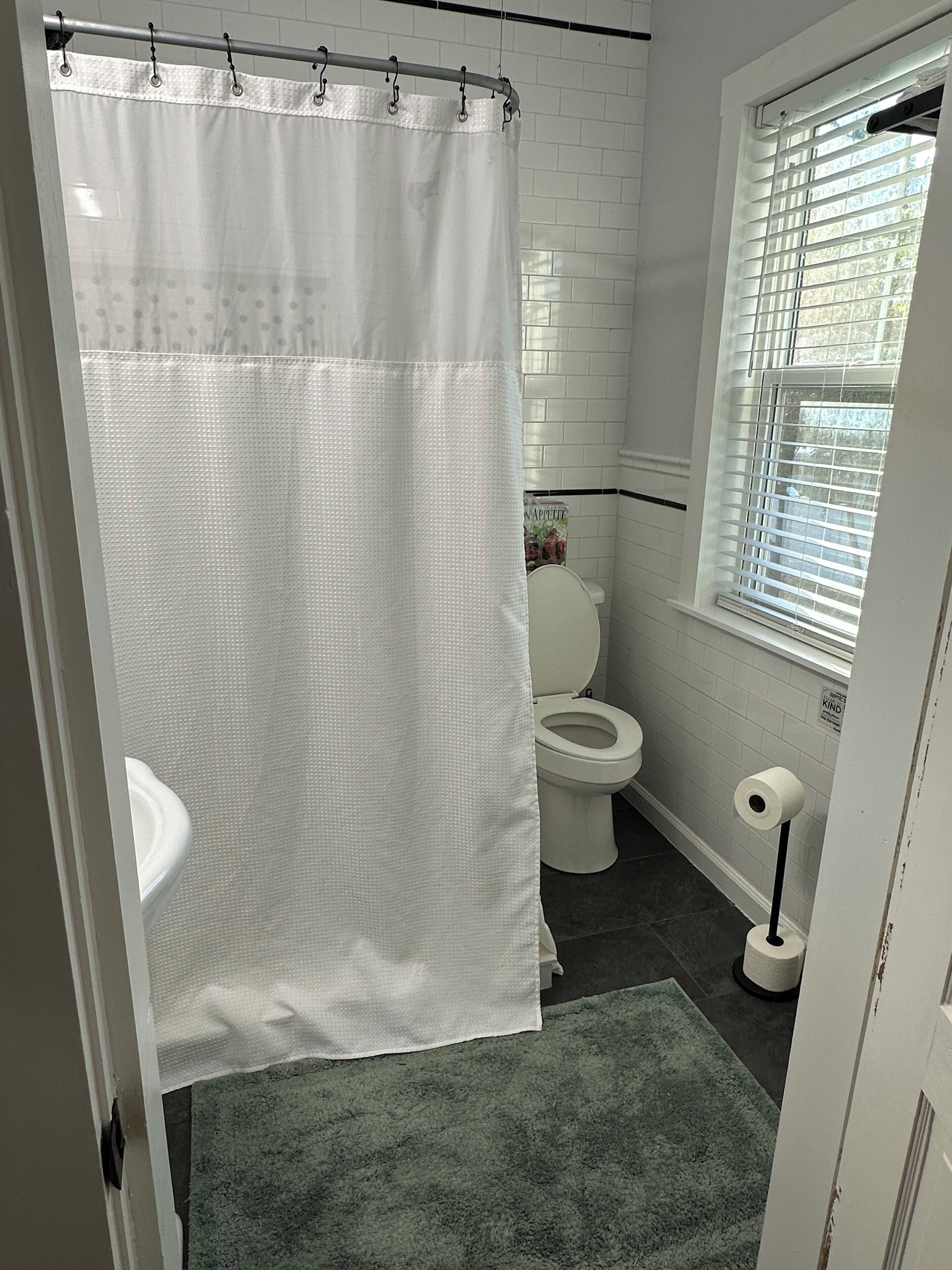 Un baño con inodoro blanco, cortina de ducha blanca, alfombra verde y una ventana con persianas.