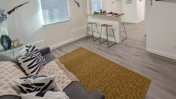 A modern apartment with a grey sofa, a patterned rug, and a kitchen area with bar stools.
