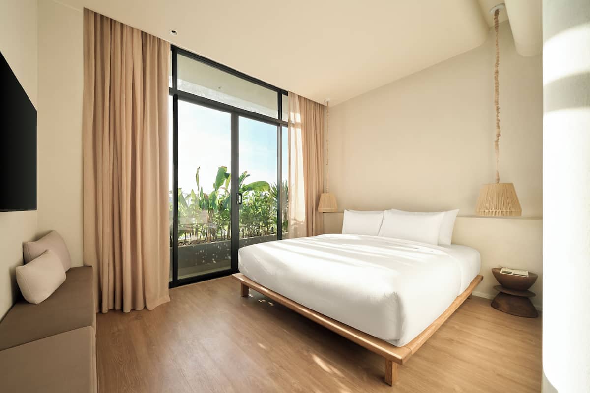 A modern hotel room with a large bed, a sofa, and a view of greenery through the window.