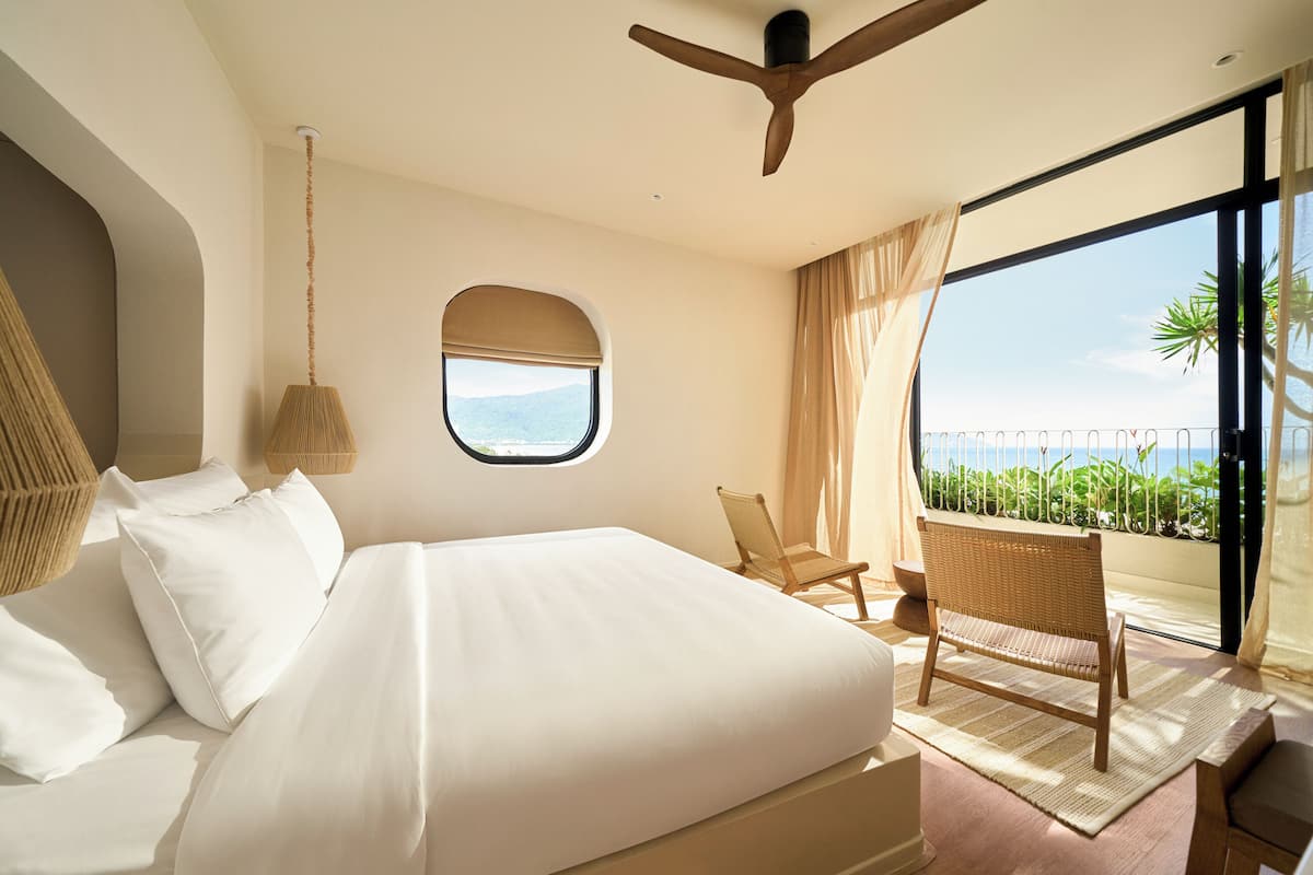 A bedroom with a large bed, a window with a view, a ceiling fan, and a chair by the window.