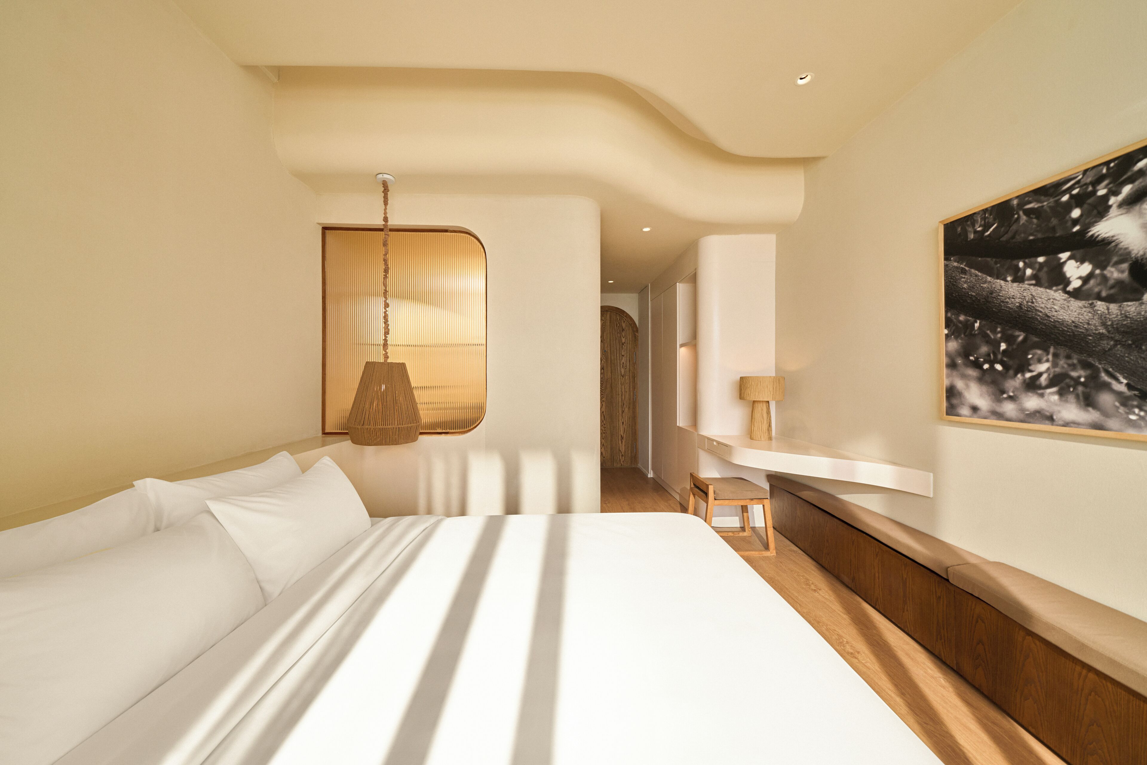 A modern hotel room with a large bed, a wooden bench, a hanging lamp, and a framed picture on the wall.