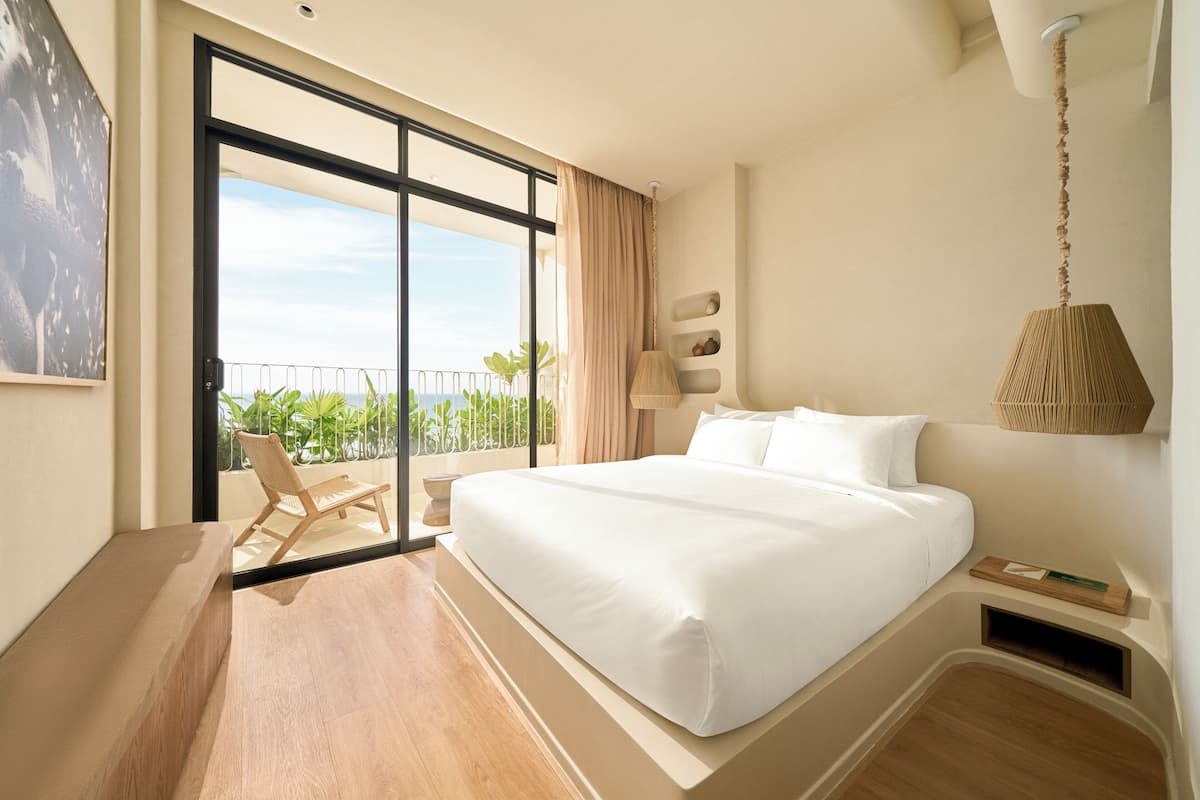 A modern hotel room with a large bed, a balcony with a chair, and a view of greenery.