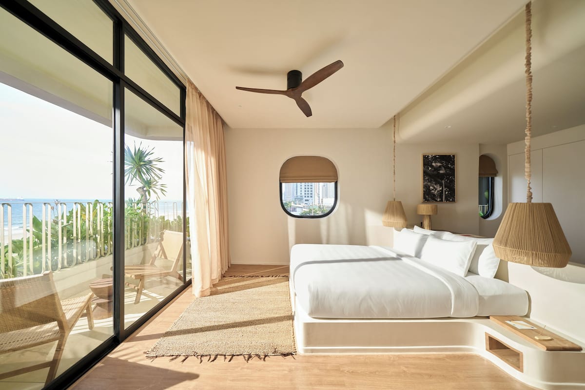 A bedroom with a large bed, a ceiling fan, a window with a view, and a balcony with chairs and a table.