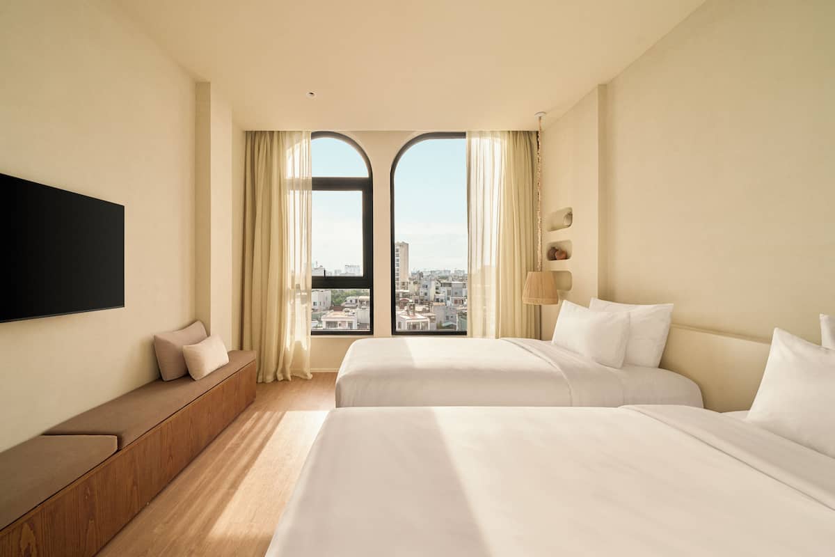 A hotel room with two beds, a sofa bed, a TV mounted on the wall, and a view of the city through large windows.