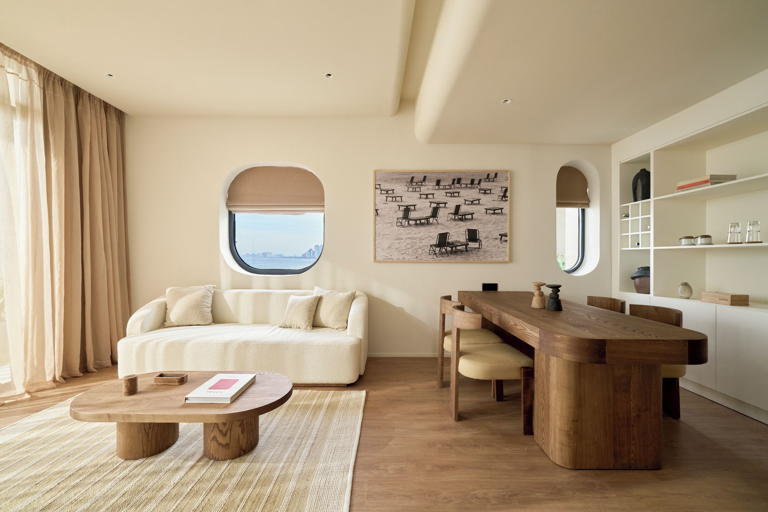 A modern living room with a sofa, coffee table, dining table, and a view through a window.