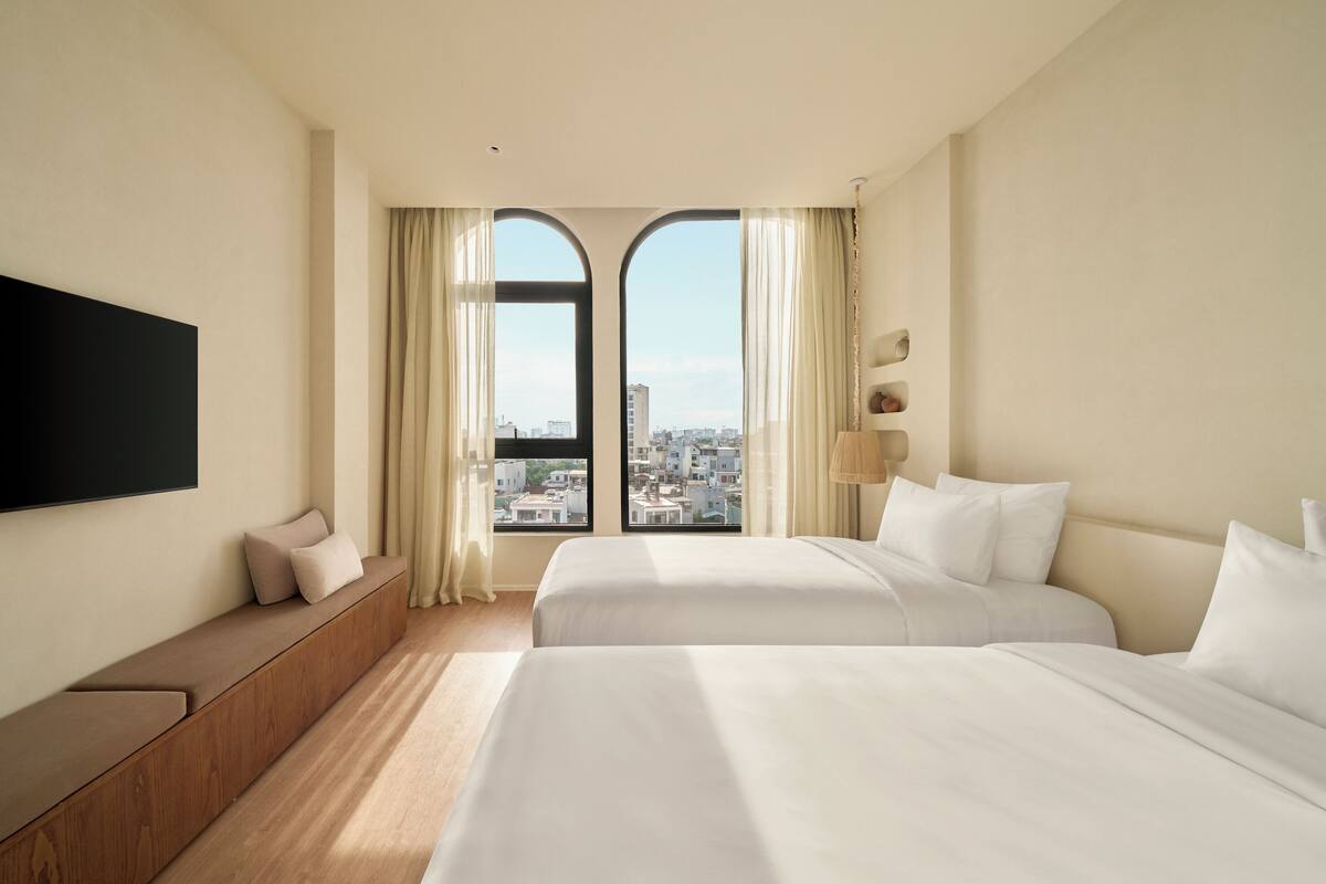 A hotel room with two beds, a sofa, a TV, and a city view through large windows.