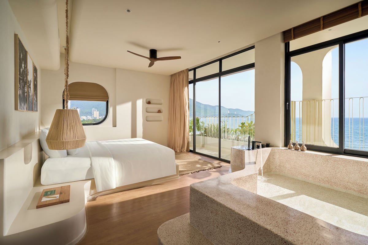A modern hotel room with a large bed, a sitting area with a view, a ceiling fan, and a large window overlooking the sea.