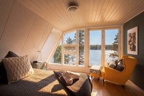 A bedroom with a large window, a bed, a chair, and a view of a lake.