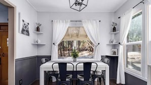 A dining area with a white table and black chairs, a window with white curtains, and shelves with decorative items.