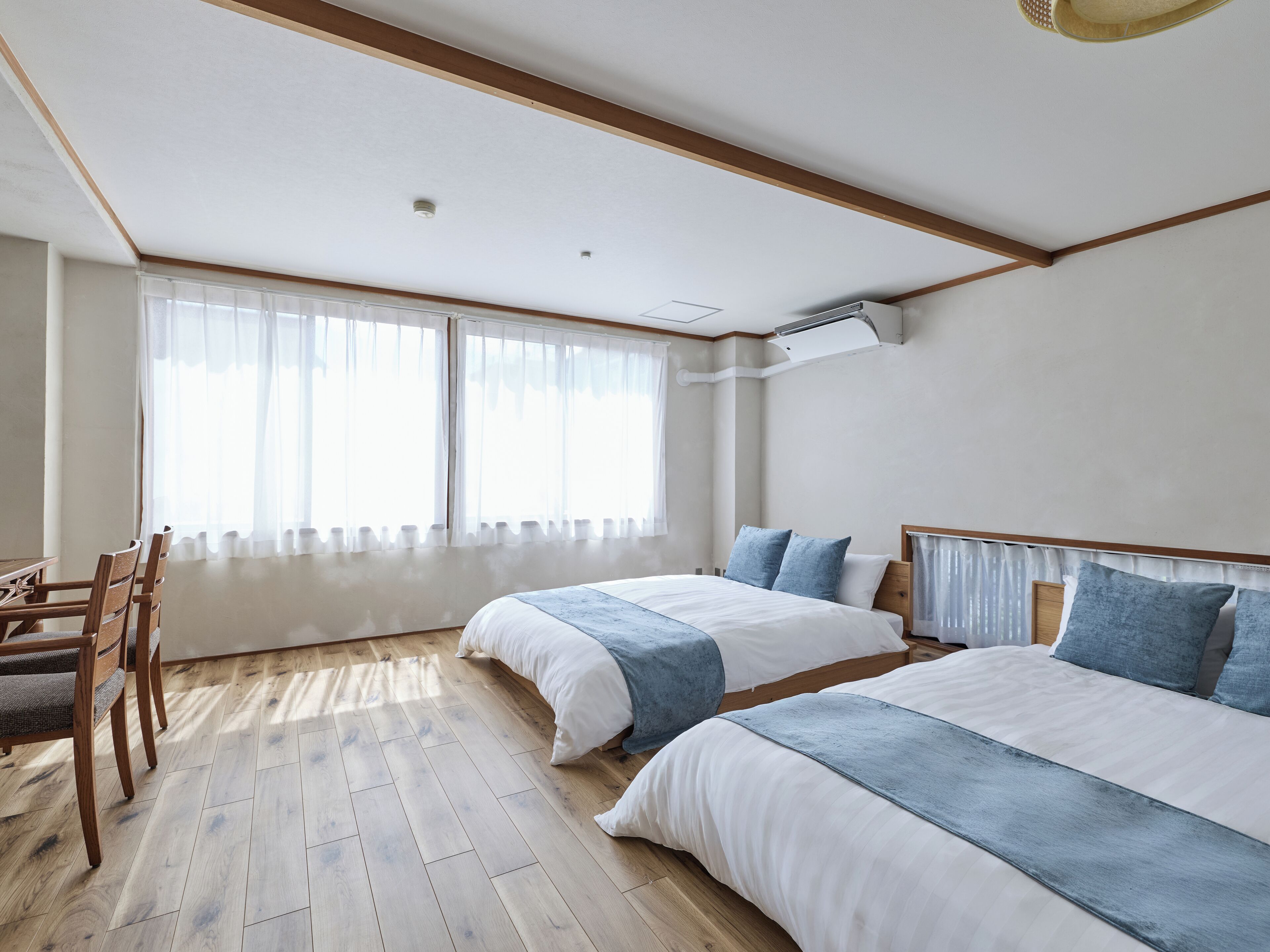 A hotel room with two beds, a wooden floor, a desk with chairs, and large windows with sheer curtains.