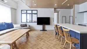 Un espai de vida modern amb terra de fusta en disseny de guilló, un gran finestral, un televisor de pantalla plana, una zona de menjador amb cadires de fusta i una cuina amb armaris blancs.