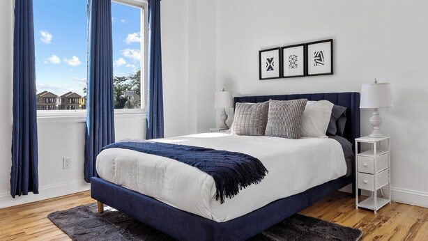 A bedroom with a large bed, a view of houses through the window, and a nightstand with a lamp.