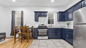 A modern kitchen with dark blue cabinets, a stainless steel refrigerator, a built-in oven, and a dining area with wooden chairs and a table.