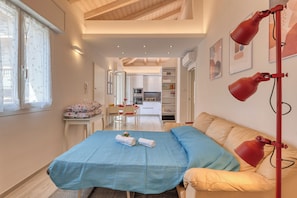 A bedroom with a bed, a desk, and a kitchenette in the background.