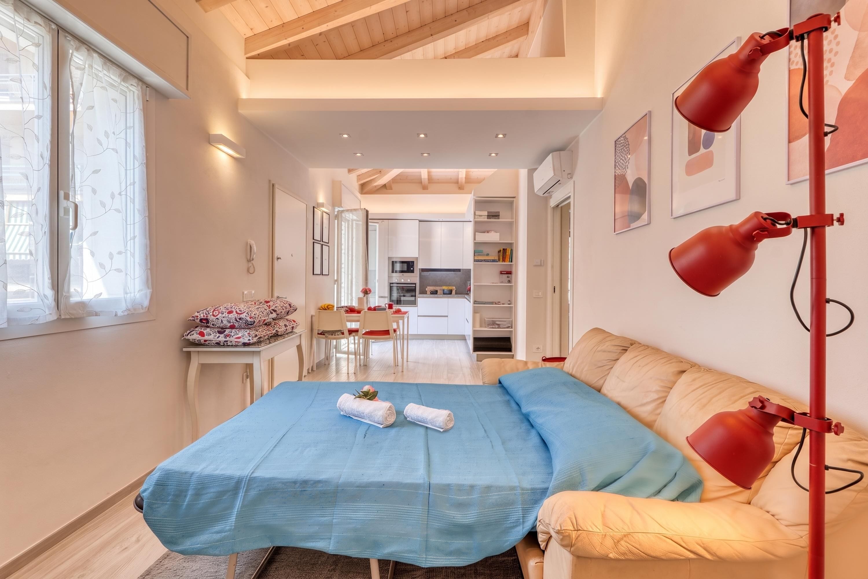 Een slaapkamer met een bed, een bureau, een stoel, een raam met gordijnen, een rode vloerlamp en een keuken op de achtergrond.