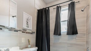 A modern bathroom with a white sink, toilet, and bathtub. There is a window with black curtains, a white cabinet, and a small vase with flowers on the counter.