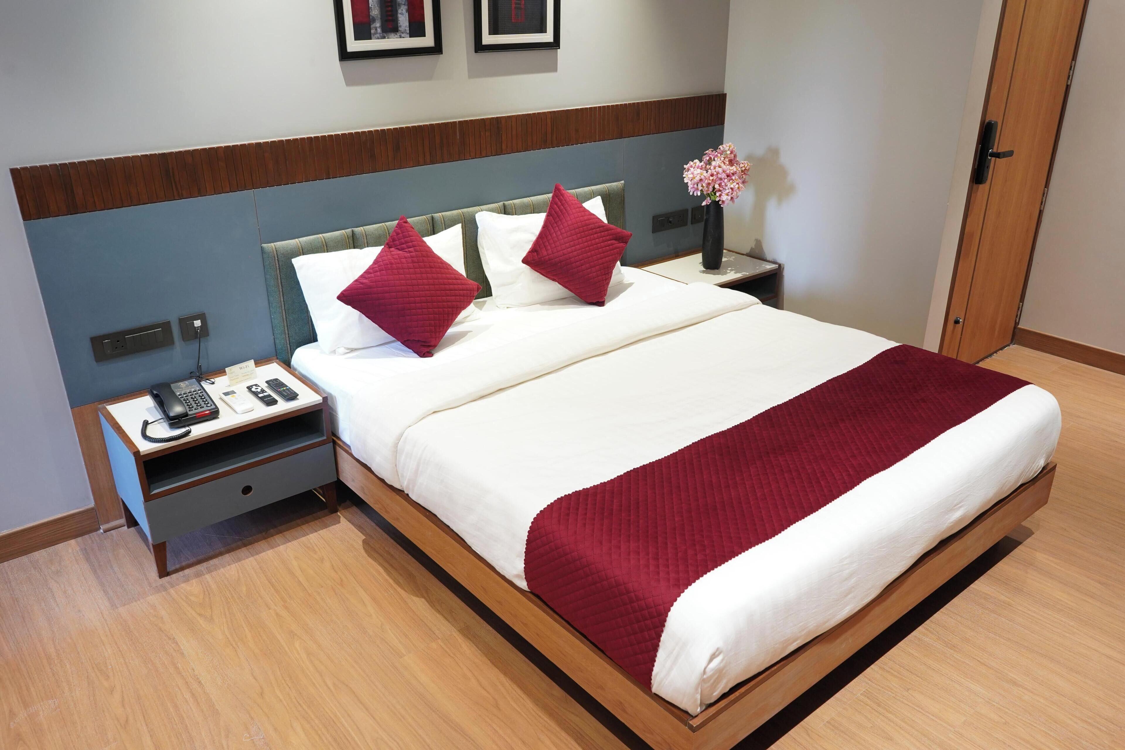 Una cama bien hecha con una colcha blanca y roja, dos almohadas rojas y un cabecero de madera. Hay una mesita de noche con un teléfono y un jarrón con flores al lado de la cama.