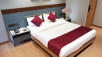 Una cama bien hecha con una colcha blanca y roja, dos almohadas rojas y un cabecero de madera. Hay una mesita de noche con un teléfono y un jarrón con flores al lado de la cama.
