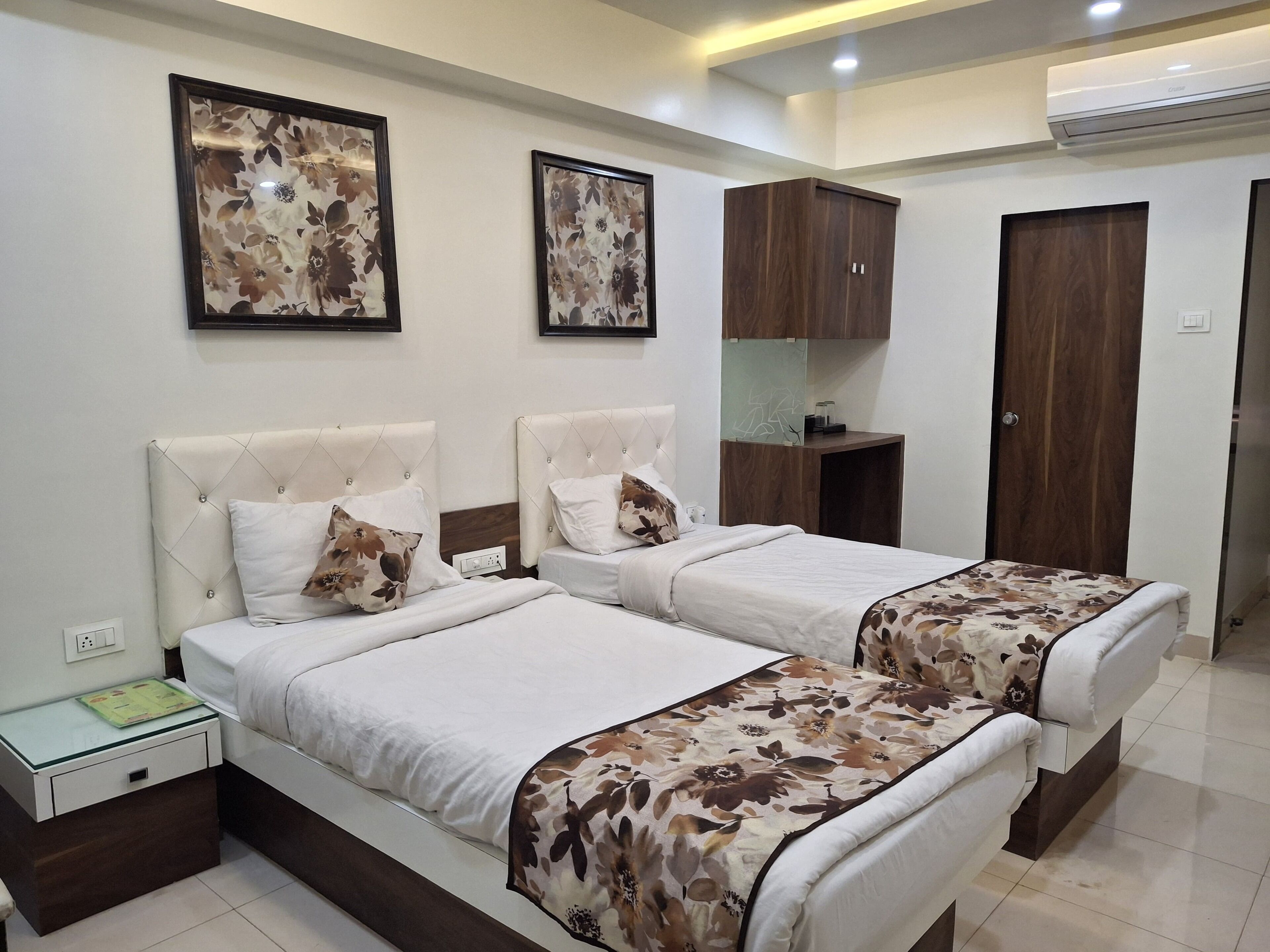A hotel room with two beds, a nightstand, a small table, a wardrobe, and framed floral artwork on the walls.