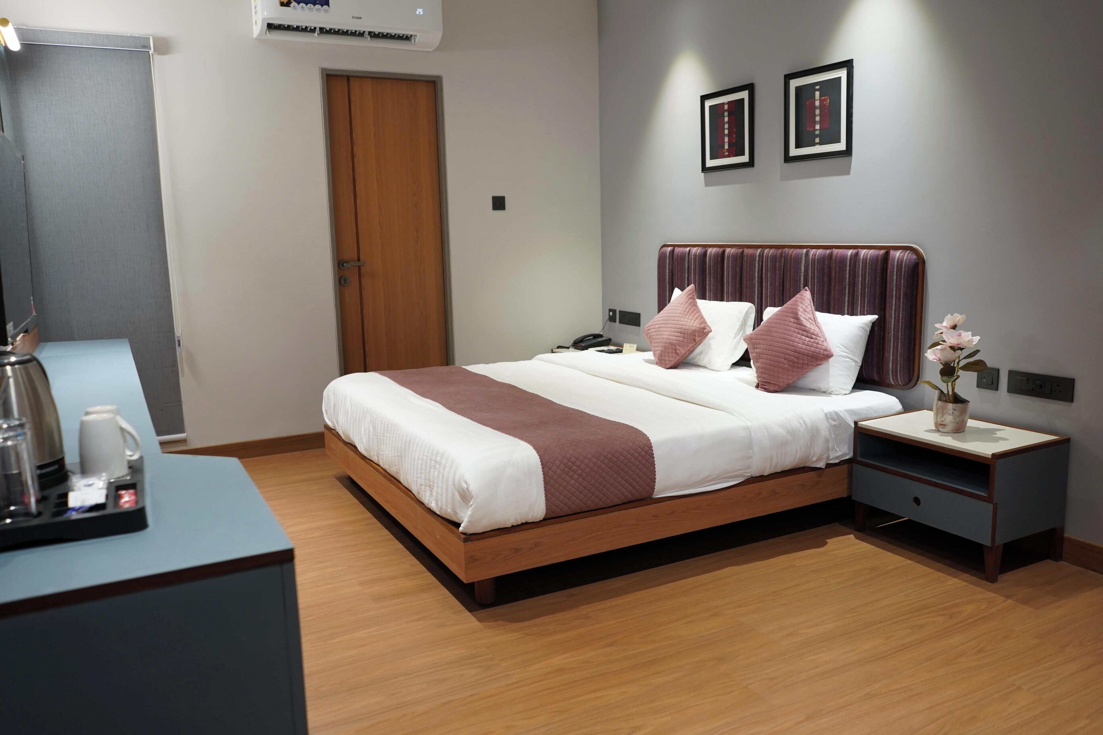 A hotel room with a wooden bed, a bedside table with a vase, and a desk with a coffee maker.