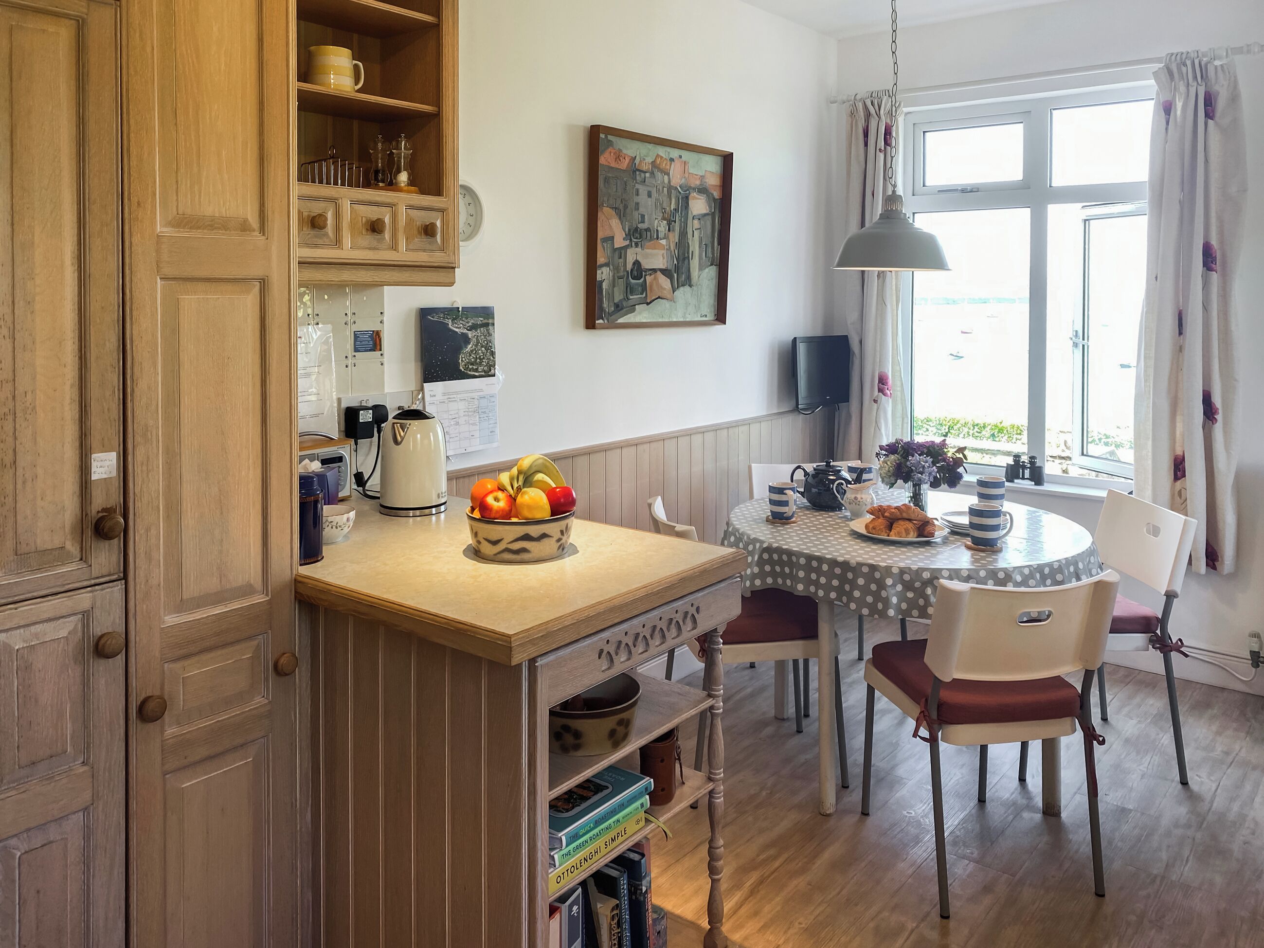 Une cuisine avec des meubles de rangement en bois, un saladier rempli de fruits posé sur le comptoir, une table avec des chaises et une fenêtre avec des rideaux.