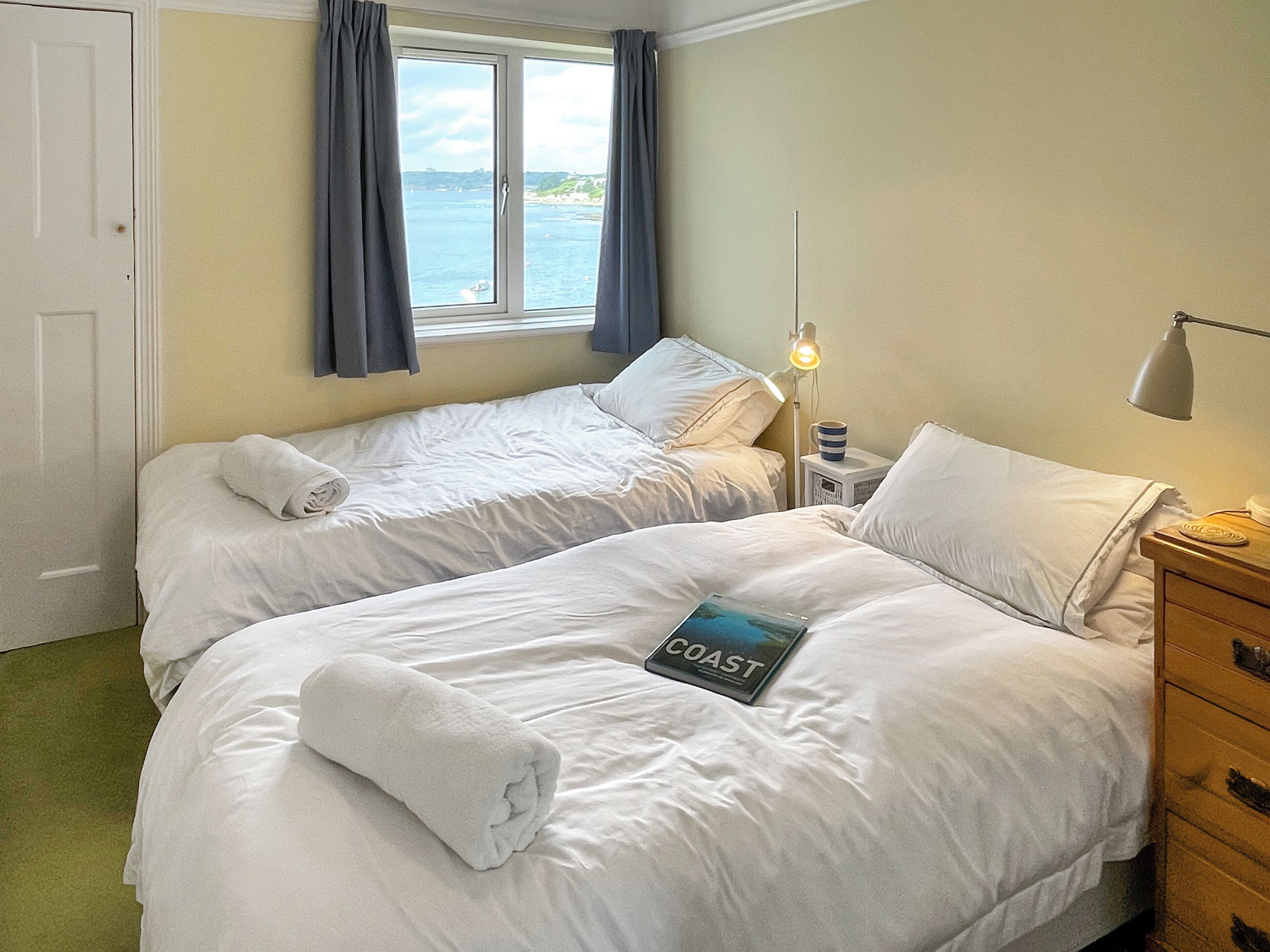 A hotel room with two beds, a nightstand with a lamp, a window with curtains, and a view of the sea.